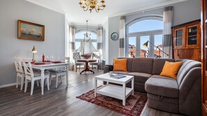 A modern living room with a dining area, featuring a chandelier, a sofa with orange cushions, and a large window offering a view of the city. - App. Lolita - Lolita (Kühlungsborn)