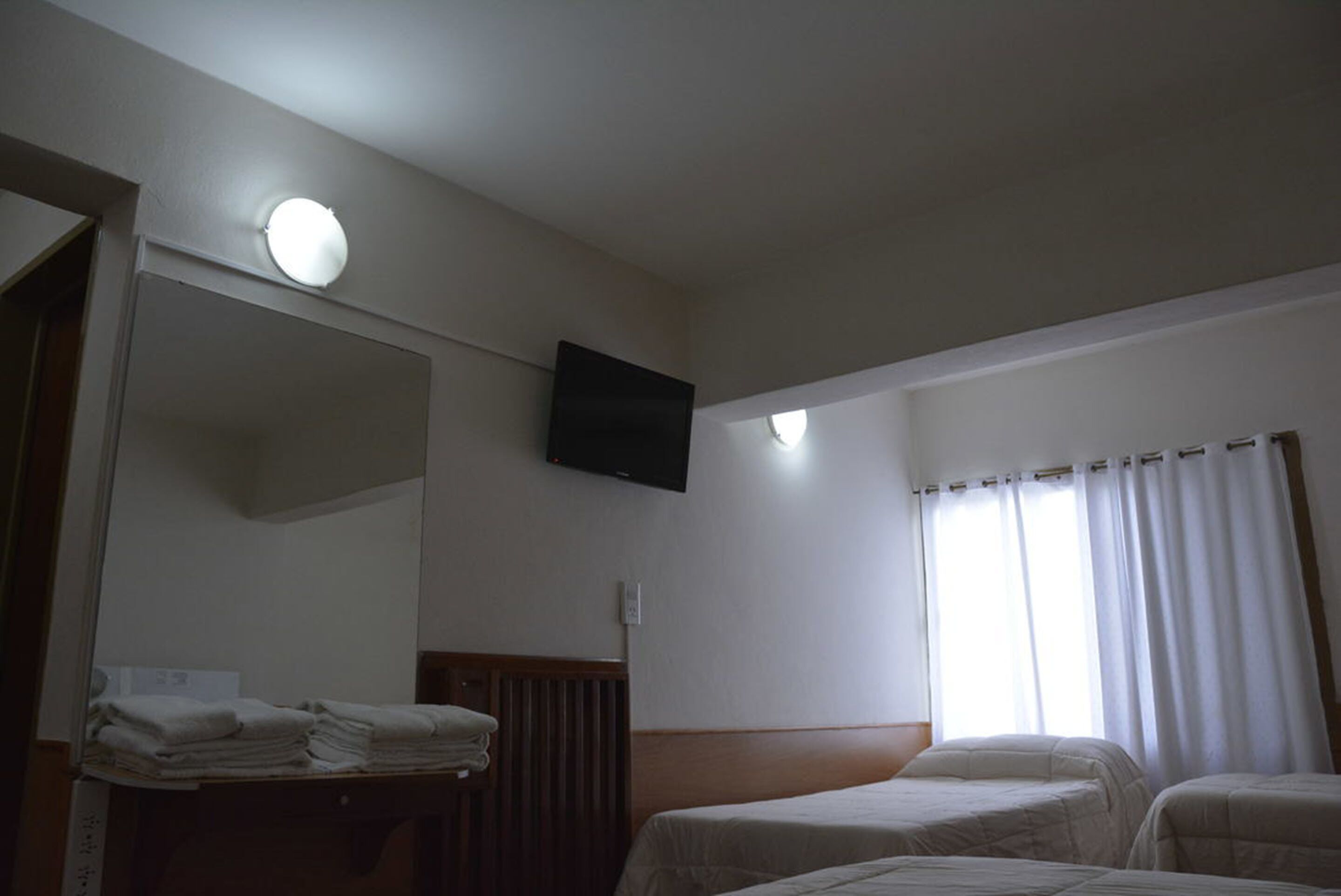 Une chambre d’hôtel avec deux lits, un téléviseur à écran plat fixé au mur, une fenêtre avec des rideaux et une pile de serviettes sur une table en bois.