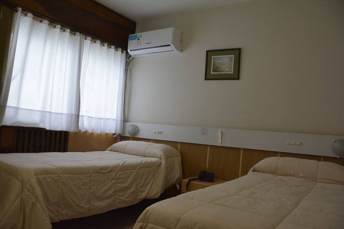 Habitación de hotel con dos camas, una ventana con cortinas, un equipo de aire acondicionado, una mesita de noche y un cuadro enmarcado en la pared.