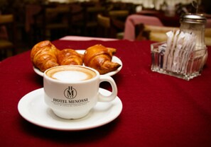 A white cup with "HOTEL MENOSSI" logo on a red tablecloth, a plate with croissants, and a glass jar with napkins. - Hotel Menossi (Rio Cuarto)