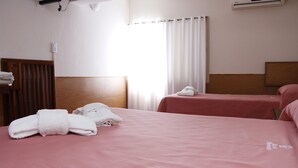 A hotel room with two beds, each with a pink bedspread and white towels. There is a window with white curtains, an air conditioning unit on the wall, and a wooden headboard. - Hotel Menossi (Rio Cuarto)