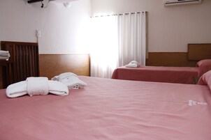Habitación de hotel con dos camas, cada una con cubrecama rosa y toallas blancas. Se ve una ventana con cortinas blancas, un equipo de aire acondicionado en la pared y un cabecero de madera.