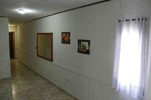 Un pasillo con un espejo grande, dos cuadros enmarcados y una ventana con cortinas blancas.