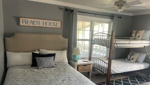 Un dormitorio con una cama, una mesita de noche, una lámpara, un ventanal con cortinas y una litera.