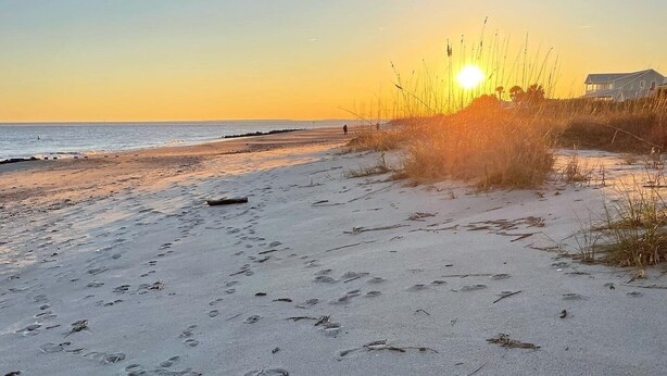 Una playa con huellas y hierba alta en primer plano, una casa al fondo y el sol poniéndose sobre el océano.