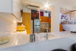 Una cocina moderna con encimeras blancas, un refrigerador de acero inoxidable y un lavamanos con grifo moderno. Hay elementos decorativos sobre la encimera y obras de arte en las paredes.