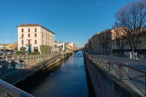 A canal flanked by buildings with balconies, a bridge overhead, and a clear sky.