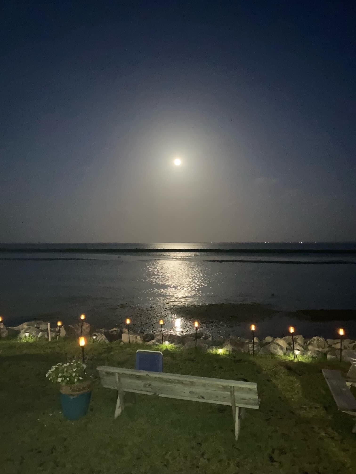 Un banc donnant sur une plage la nuit, avec une mer illuminée par la lune et des lanternes en pierre.