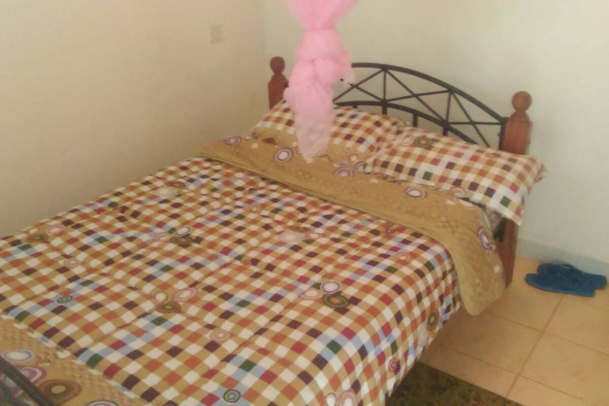 A single bed with a checkered bedspread and pillows, a wooden headboard, and a pair of shoes on the floor.
