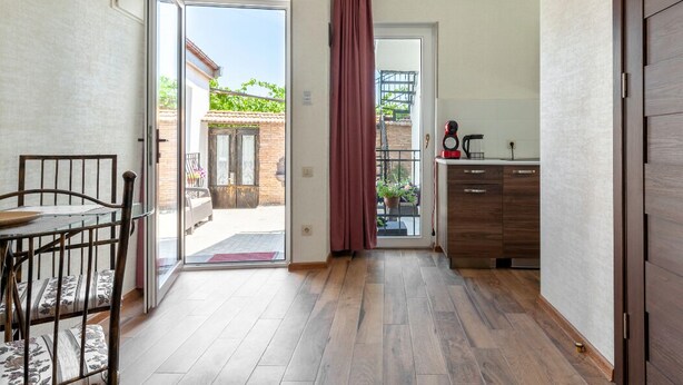 A spacious room with a wooden floor, a dining area with a table and chairs, a kitchen with wooden cabinets, and a sliding glass door leading to an outdoor area with a patio and plants. - Studio Apartment on top of old Tbilisi with a 180 degree View of the city (Dzveli Tbilisi)