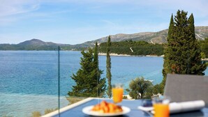 A table with a plate of food and a glass of orange juice, overlooking a body of water and hills with wind turbines. - Hedera Estate, Villa Hedera XV (Slano)