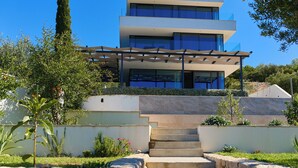 A modern multi-story building with a glass facade, a stone staircase leading to the entrance, and a well-maintained garden with various plants and a tall tree. - Hedera Estate, Villa Hedera XV (Slano)