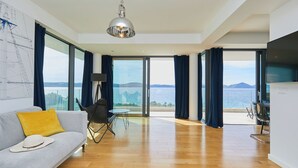 A spacious living room with a large window, a sofa with a yellow pillow, a dining area, and a ceiling light. - Hedera Estate, Villa Hedera XV (Slano)