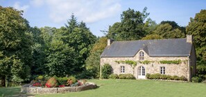 A stone house with a sloped roof, surrounded by lush greenery and a well-maintained lawn.
