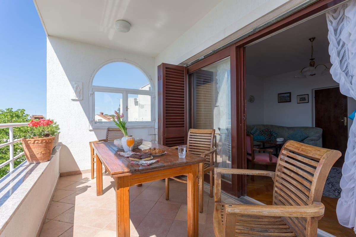 A balcony with a wooden table and chairs, a potted plant, and a view of the interior.