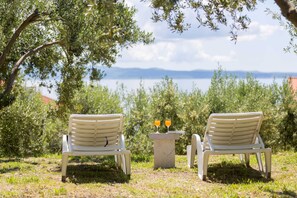 Two white chairs and a small table with two glasses of orange juice in a grassy outdoor area with trees and a view of a body of water. - Apartment Sunrise (Split)