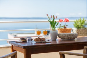 Una mesa de madera con croissants, jugo de naranja y un florero con flores en un balcón con vistas al mar.