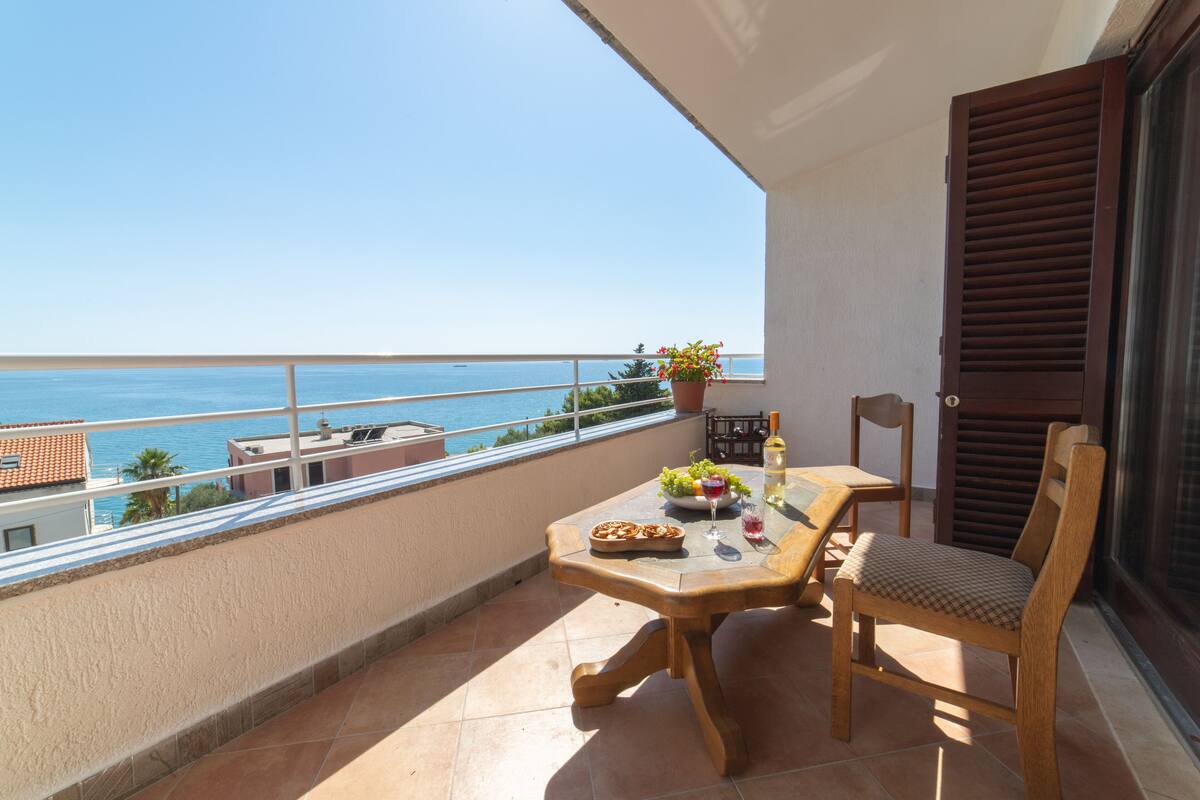 A balcony with a table set for two, a chair, a potted plant, and a view of the sea.
