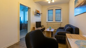 Ein gemütliches Wohnzimmer mit einer Couch, einem kleinen Tisch und einem Fernseher. Ein Fenster mit Jalousien und eine Tür, die in einen anderen Raum führt.