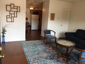 Un salon avec un canapé foncé, deux fauteuils bleus, une table basse ronde et un tapis à motifs. Un vase bleu contenant des plantes se trouve à gauche, et une décoration murale en bois est accrochée au mur.