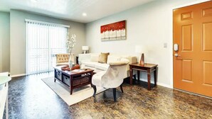 A living room with a sofa, coffee table, and lamps. There is a door and a window with blinds. - Evie's Campus-close 2 bedroom luxury (Norman)