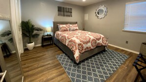 A bedroom with a bed, a nightstand, a lamp, a potted plant, a mirror, and a window with blinds. - Evie's Campus-close 2 bedroom luxury (Norman)