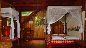 Uma cabana de madeira com duas camas, cada uma com dossel. Há uma cadeira de madeira e uma pequena mesa.