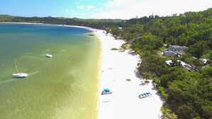 Uma praia com águas turquesa cristalinas, areal arenoso e alguns barcos. Há casas e árvores ao longo da praia.