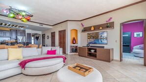 Una sala de estar moderna con un sofá, reposapiés y una bandeja con aperitivos. Se observa un televisor, una chimenea y una zona de cocina al fondo.