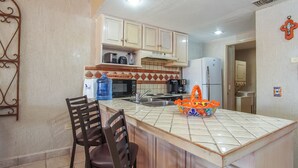 A compact kitchen with a central island, a sink, and a refrigerator. There are two chairs and a large water dispenser. - Princesa Resort D-509 Whispering Sands Great Ocean View Condo (Puerto Peñasco)