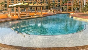 A large, clear swimming pool with a circular design, surrounded by palm trees and a multi-story hotel building in the background. - Princesa Resort D-509 Whispering Sands Great Ocean View Condo (Puerto Peñasco)