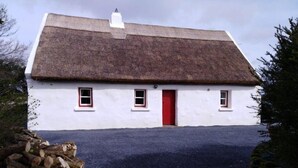 Une maison blanche avec un toit de chaume, une porte rouge et deux fenêtres aux encadrements rouges.