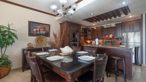 A dining area with a dark wood table, chairs, and a chandelier. There are cabinets and a refrigerator in the background.