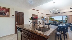 Una cocina con encimeras de granito, un fregadero de acero inoxidable y un cuenco con fruta. Hay un comedor con vista al océano.