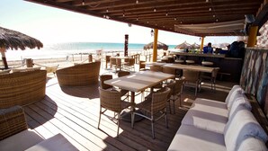 Un restaurante frente a la playa con terraza de madera, sombrillas de paja y vista al océano.