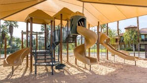 A playground with slides and climbing structures, surrounded by a sandy area and palm trees.