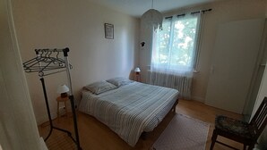 A bedroom with a bed, a radiator, a window with curtains, a chair, and a hanging clothes rack.