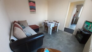 A living room with a black leather sofa, a small round table with a basket and a bottle, a dining table with chairs, and a TV on a stand.