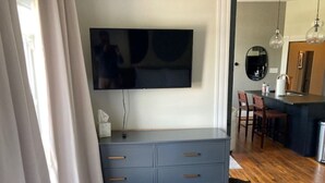 A room with a flat-screen TV mounted on the wall, a grey dresser with drawers, a wooden floor, and a kitchen area with a sink and chairs. - Modern Luxe Apartment - Close to City Center (Richmond)