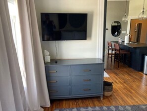Une pièce dotée d’un téléviseur à écran plat fixé au mur, d’une commode grise avec des tiroirs, d’un plancher en bois et d’un coin cuisine avec un évier et des chaises.