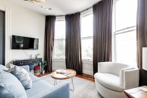 Ein modernes Wohnzimmer mit einem wandmontierten Flachbildfernseher, einem runden Holztisch, einer Couch, einem Sessel und einem großen Fenster mit Vorhängen.