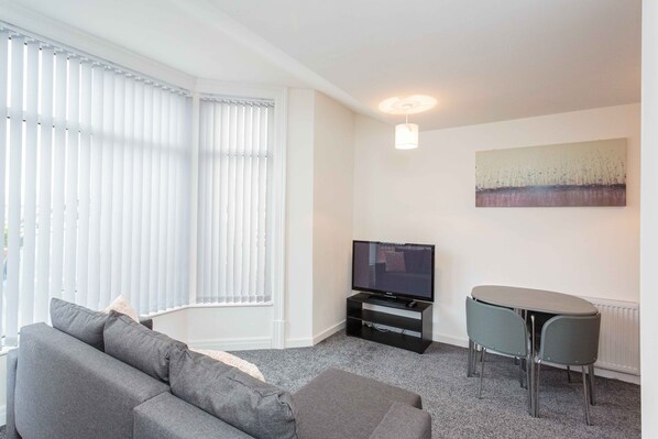 A living room with a grey sofa, a TV on a black stand, a small table with two chairs, and a painting on the wall. - Cherry Property - Hornby Road (Blackpool)