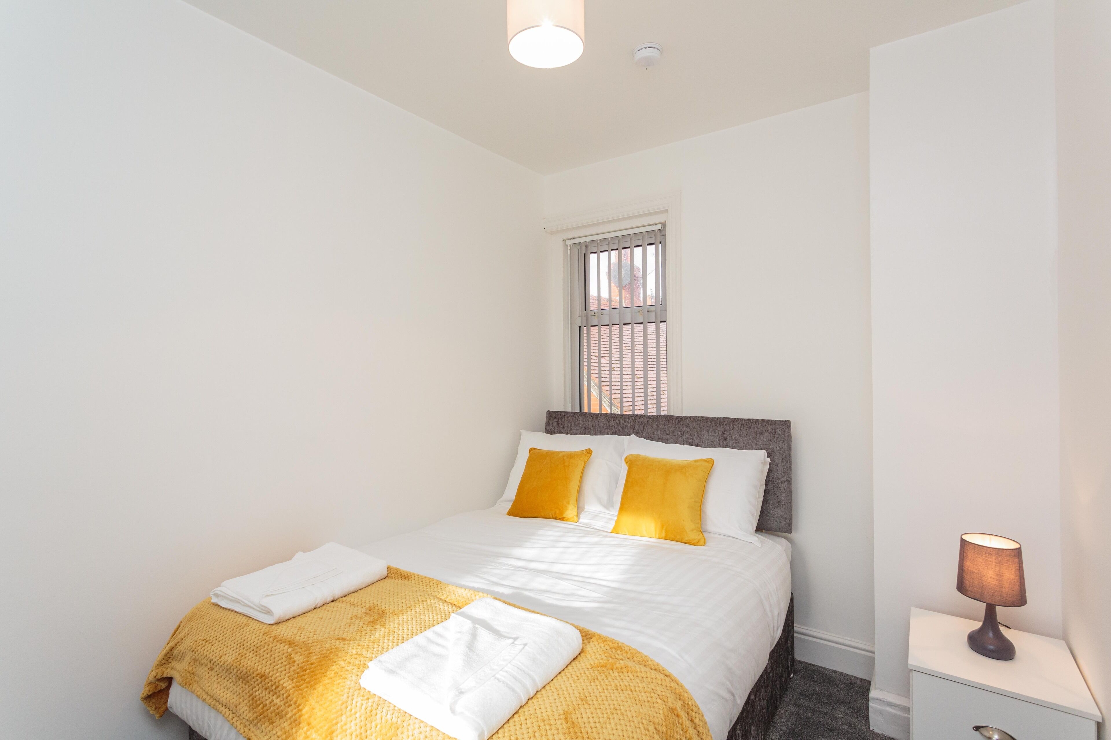 A neatly made bed with white and yellow bedding, a bedside lamp, and a window with blinds.