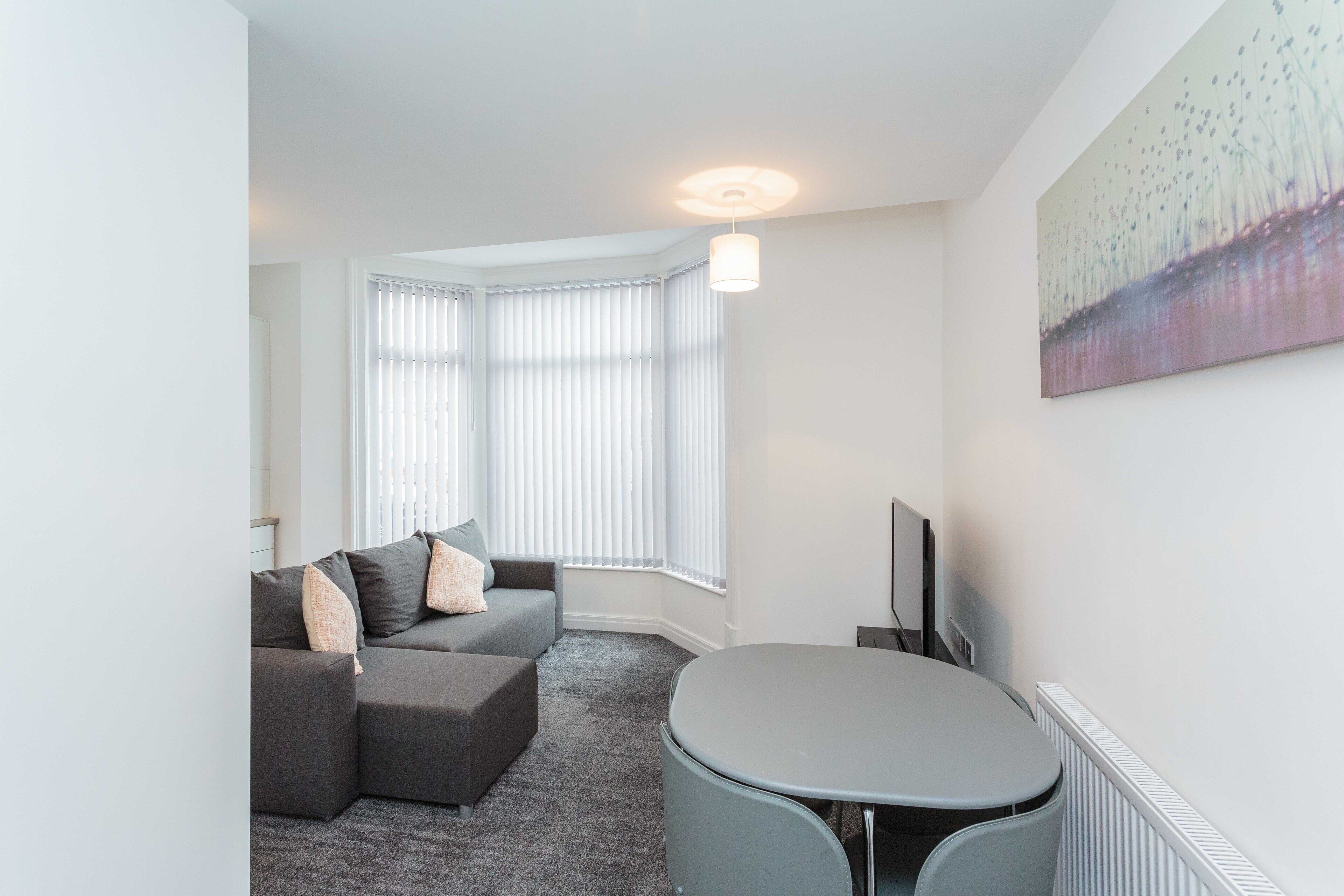 A modern living room with a grey sectional sofa, a round table, and a large window with blinds.