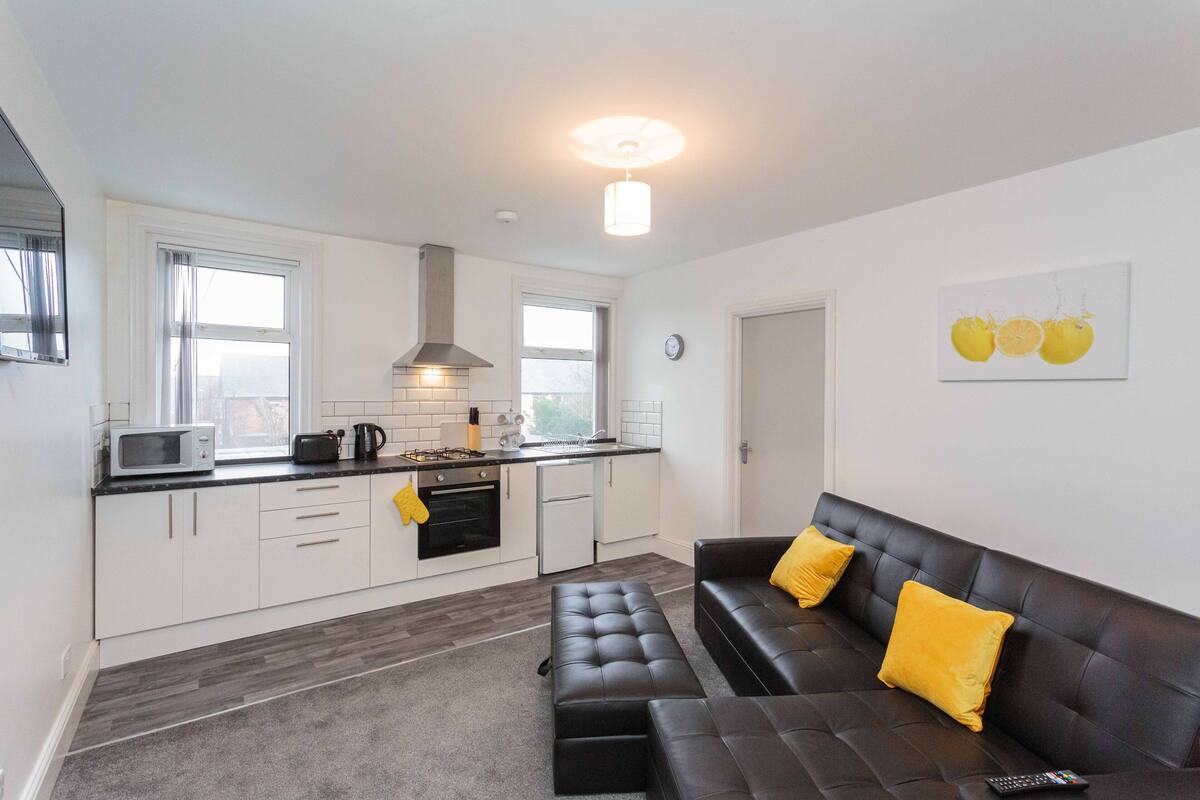 A modern kitchen with white cabinets, a black countertop, and a black leather sofa with yellow cushions.