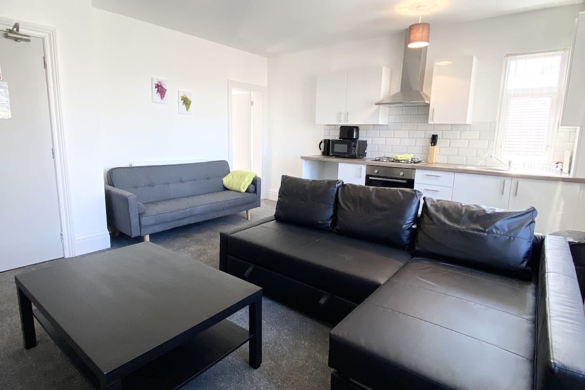 A modern living room with a grey sofa, black leather ottoman, and a black coffee table. A kitchen with white cabinets and a black microwave is visible in the background.