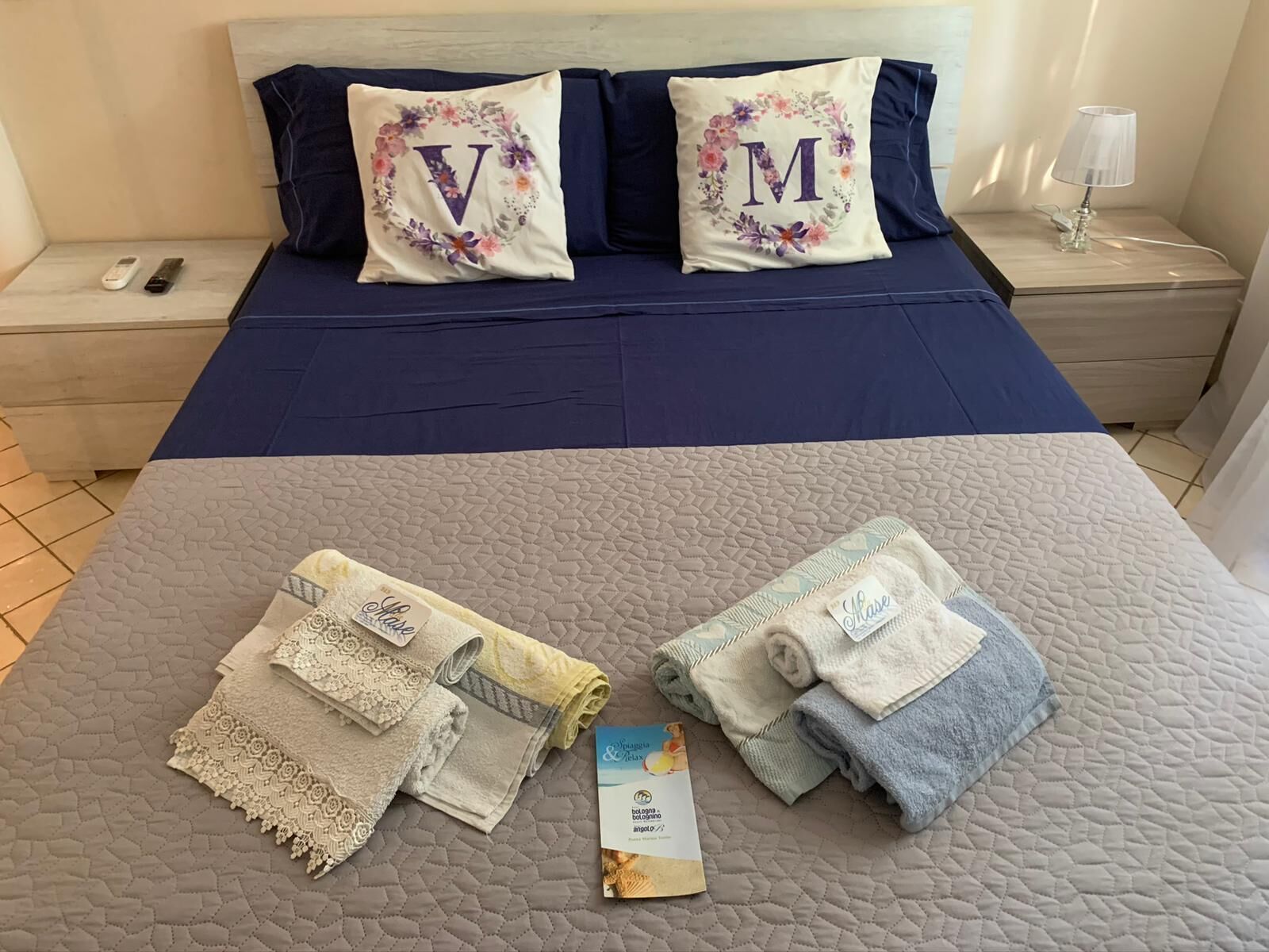 Een net opgemaakt bed met twee kussens met bloemenmotieven en grote letters, een nachtkastje met een lamp, en een opgevouwen handdoek met een label.