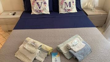Een net opgemaakt bed met twee kussens met bloemenmotieven en grote letters, een nachtkastje met een lamp, en een opgevouwen handdoek met een label.