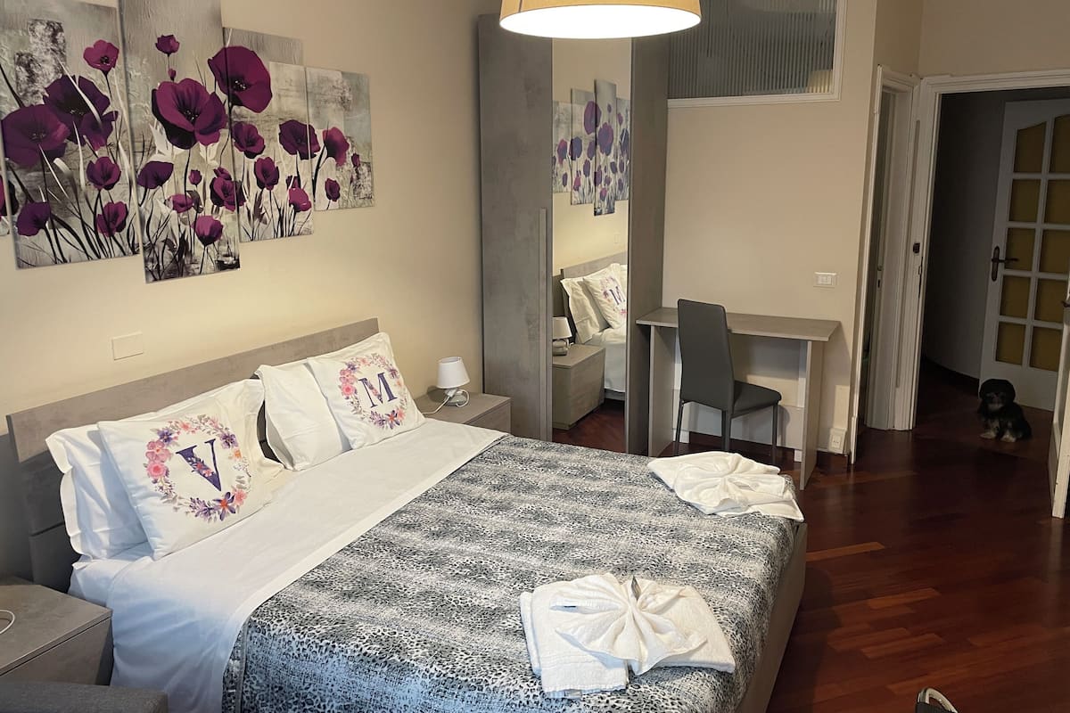 A bedroom with a bed, a desk, and a chair. There are floral paintings on the wall.