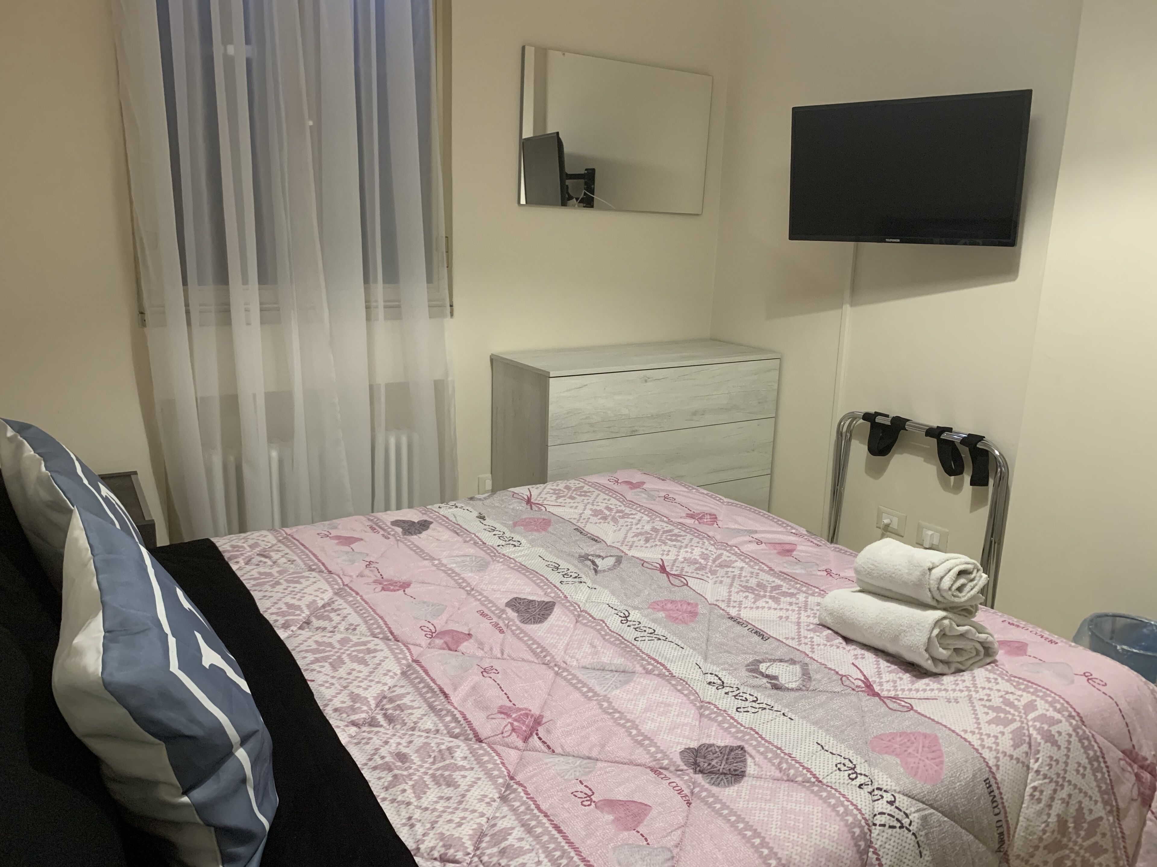 A hotel room with a bed, a bedside table, a dresser, a TV mounted on the wall, and a window with sheer curtains.